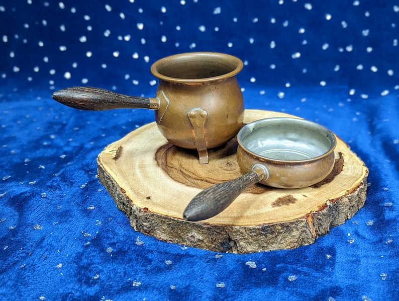 May include: Two copper pots with wooden handles rest on a wooden slice against a blue background. The larger pot has a rounded body and a small spout. The smaller pot is a shallow bowl shape. Both pots have a vintage aesthetic.