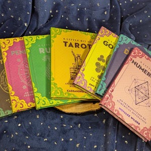 May include: A collection of colorful books with titles like "A Little Bit of Tarot" and "Numerology." The books have decorative borders and feature illustrations and text. The books are arranged on a dark blue, textured surface.
