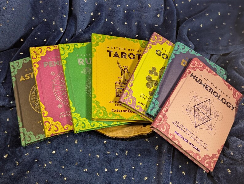 May include: A collection of colorful books with titles like "A Little Bit of Tarot" and "Numerology." The books have decorative borders and feature illustrations and text. The books are arranged on a dark blue, textured surface.