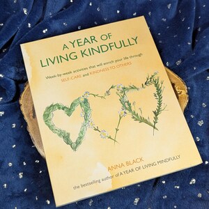 May include: A hardcover book titled "A Year of Living Kindly" with a light yellow cover. The cover features a heart and a stylized "&" symbol made of green leaves and white flowers. The book is by Anna Black.