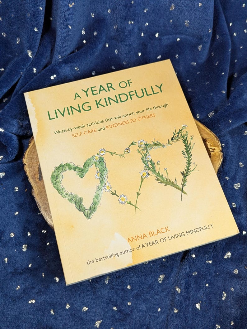 May include: A hardcover book titled "A Year of Living Kindly" with a light yellow cover. The cover features a heart and a stylized "&" symbol made of green leaves and white flowers. The book is by Anna Black.