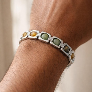 Könnte beinhalten: Silberarmband mit ovalen Edelsteinen in Gelb, Grün und Braun. Das Armband hat ein rechteckiges Kettendesign mit kleinen, klaren Steinen zwischen jedem Edelstein. Das Armband wird am Handgelenk getragen.