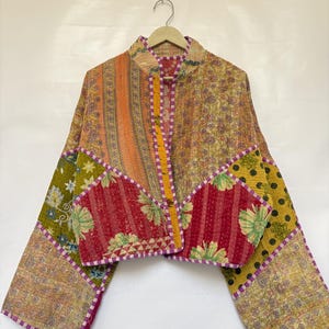 May include: A cropped jacket made from quilted patchwork fabric featuring a variety of floral, paisley, and polka dot patterns in shades of yellow, orange, red, green, and purple. The garment has long sleeves and a purple and white striped trim.