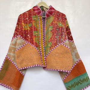 May include: A cropped jacket made from quilted fabric featuring a patchwork of red, orange, green, and yellow patterns. The garment has long, wide sleeves, a stand-up collar, and two large front pockets, all finished with purple and white striped trim.