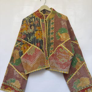 May include: A cropped jacket made from quilted fabric featuring a patchwork of floral and botanical patterns in shades of brown, yellow, green, and red. The garment has long sleeves, a mandarin collar, and yellow and white striped trim.