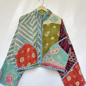 May include: A cropped jacket constructed from various patchwork fabric panels featuring floral, striped, and geometric patterns in shades of green, red, blue, and cream. The garment has a mandarin collar and blue and white striped trim.