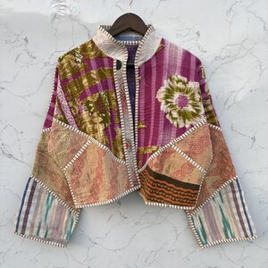May include: A cropped jacket made from quilted patchwork fabric featuring a mix of floral and geometric patterns in shades of magenta, olive green, and cream. The garment has a high collar, long sleeves, and visible white running stitch details.