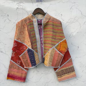 Könnte beinhalten: Eine kurz geschnittene Jacke aus Patchwork-Stoff mit einer Vielzahl bunter Muster, darunter Streifen, florale Motive und geometrische Formen in Rot-, Orange-, Gelb- und Violetttönen, abgerundet mit weiß abgesteppten Kanten.