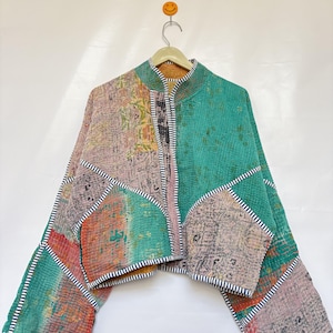 May include: A cropped jacket featuring a patchwork design of green, pink, and tan fabric panels with visible stitching. The garment has long sleeves, a stand-up collar, and is finished with black and white striped trim along the edges.