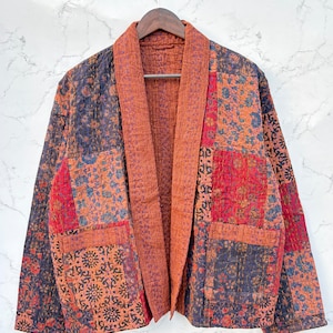 May include: A quilted jacket featuring a patchwork design of various floral and geometric patterns in shades of red, orange, blue, and black. The garment has a solid burnt orange collar, long sleeves, and two front patch pockets with visible stitching.