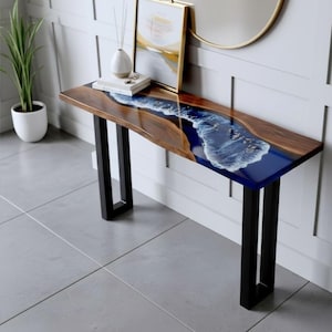 Puede incluir: Una consola de madera con un centro de resina epoxi azul en forma de río. La mesa tiene un borde de madera oscura y patas de metal negro. Una foto enmarcada, un jarrón y libros están sobre la mesa. La mesa mide aproximadamente 122 cm de largo.