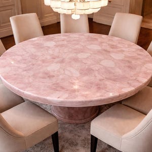 Puede incluir: Una mesa de comedor redonda con una superficie rosa estampada está rodeada de ocho sillas color crema. La mesa tiene una base de pedestal y está iluminada por una lámpara de araña. Las sillas tienen patas oscuras y respaldos curvos.