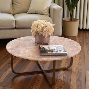 May include: A circular coffee table with a pink marble effect top and a gold-coloured metal frame. A vase of dried flowers and a book rest on the table. The table is in a living room.
