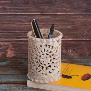 Puede incluir: Un portalápices de piedra beige tallada con un diseño floral, que contiene un bolígrafo negro y dorado y un bolígrafo plateado. El portalápices está sobre un libro amarillo con un círculo rojo en la portada, sobre un fondo de madera.