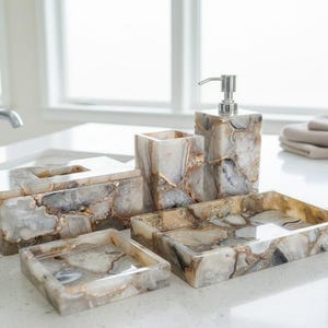 Puede incluir: Un juego de baño que incluye una caja de pañuelos, un dispensador de jabón, un vaso y dos bandejas. El juego está hecho de un material similar a la piedra con un patrón veteado en tonos grises, blancos y dorados. El dispensador de jabón tiene una bomba plateada.