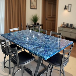 Op de afbeelding: Een rechthoekige eettafel met een blauwe agaat stenen blad. De tafel wordt ondersteund door een zilverkleurig metalen onderstel en is omringd door zes stoelen met zwarte zittingen en zilverkleurige metalen frames. De tafel is ongeveer 200 cm lang.