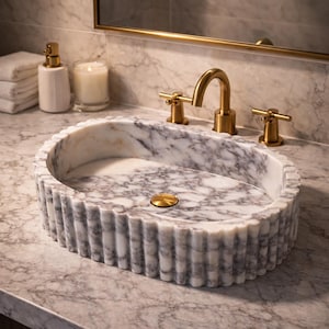 Puede incluir: Lavabo ovalado de m&aacute;rmol con exterior estriado y accesorios dorados. El lavabo est&aacute; sobre una encimera de m&aacute;rmol, con un grifo dorado y manijas a juego. Se ven un dispensador de jab&oacute;n y una vela.