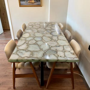 May include: A rectangular dining table with a unique, light green and white stone top. The table has wooden legs and is surrounded by four upholstered chairs. A framed picture hangs on the wall above the table.