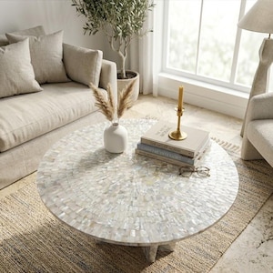 Puede incluir: Una mesa de centro redonda con una tapa de mosaico de concha es el punto focal de una sala de estar. La mesa está rodeada por un sofá beige, una alfombra tejida y una planta en maceta. Libros, una vela y gafas descansan sobre la mesa.