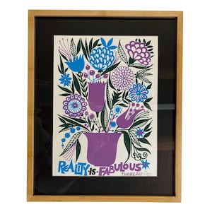 Può includere: Stampa artistica incorniciata con un motivo floreale in un vaso viola, con fiori blu, viola e bianchi e foglie verdi. Il testo "Reality is Fabulous" è stampato in blu. L'opera d'arte è incorniciata in una cornice di legno marrone chiaro.