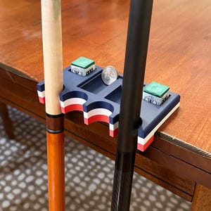 May include: A blue, white, and red pool cue holder with two cues, two green chalk cubes, and a quarter. The holder has three cue slots and a small tray. The cues are brown and black.