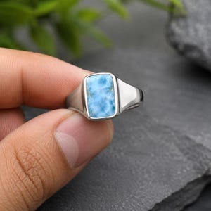 May include: A silver ring featuring a rectangular, light blue Larimar gemstone with white marbled patterns. The stone is set in a smooth, polished metal bezel, and the band has a solid, tapered design that widens toward the setting.