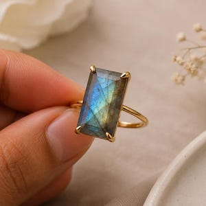 Puede incluir: Un anillo de oro con una gran piedra preciosa de labradorita rectangular facetada con destellos iridiscentes azules y amarillos. La piedra está asegurada en un engaste de cuatro garras sobre una banda de oro fina y pulida.