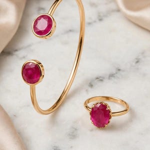 May include: A gold cuff bracelet featuring two round, faceted, deep pink rubies at the open ends, paired with a matching gold ring featuring a single oval, faceted, deep pink ruby set within a scalloped gold bezel setting on a thin band.