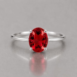 Könnte beinhalten: Ein silberfarbener Ring mit einem markanten, oval geschliffenen, leuchtend roten Edelstein, der von vier zarten Krappen gehalten wird. Die Ringschiene ist glatt und poliert und unterstreicht das schlichte Design sowie die rote Farbe.