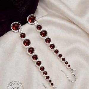 May include: A pair of long drop earrings featuring a vertical row of graduated circular dark red garnet stones set in sterling silver. Each earring is finished with a silver hook wire and includes a stamp reading STERLING 925 SILVER.