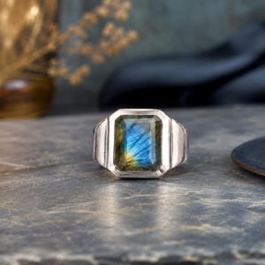 May include: A silver ring featuring a rectangular, faceted labradorite gemstone. The stone displays iridescent blue and gold flashes within its dark, semi-translucent surface. The metal band has a smooth, polished finish with a wide, structured design.