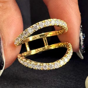 May include: A gold-colored ring with two curved bands, each encrusted with small, clear gemstones. The ring has a unique design with a connecting bar between the bands. The ring is held against a dark background with pearls.