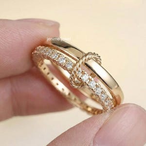 May include: A gold ring featuring a knot design. One band is set with small, clear stones. The ring has a polished finish and is held by a person's fingers. The brand name "jenime jewel" is visible.