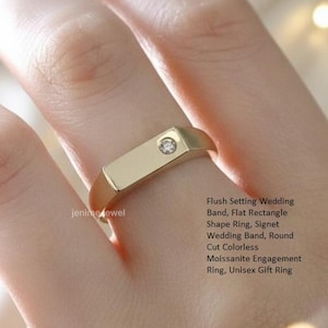 May include: A gold-colored ring with a flat rectangular shape and a small, round, colorless stone set in the center. The ring is on a finger. Text on the side reads: Flush Setting Wedding Band, Flat Rectangle Shape Ring, Signet.
