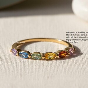 May include: A gold ring with a series of marquise-cut gemstones in rainbow colors. The ring is delicate and features a half-eternity design. Text on the side reads: Marquise Cut Wedding Band, Half Eternity Rainbow Band, Delicate, Colorfull Band, Multicolored, Engagement Band, Sapphire Dot Dash, Rainbow Band.