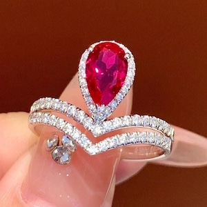 May include: A silver ring featuring a pear-shaped, vibrant red gemstone at the center, surrounded by a halo of small, clear stones. The ring has a double-band design with additional small stones, creating a sparkling, elegant appearance.