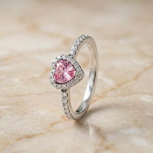 May include: A silver ring featuring a heart-shaped pink gemstone surrounded by small, clear stones. The band is also adorned with small, clear stones. The ring is set against a neutral background.