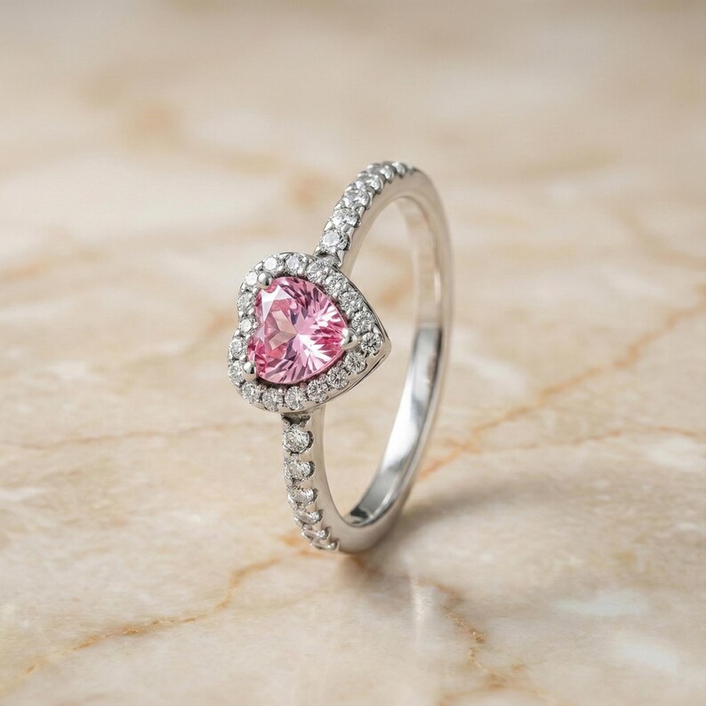 May include: A silver ring featuring a heart-shaped pink gemstone surrounded by small, clear stones. The band is also adorned with small, clear stones. The ring is set against a neutral background.