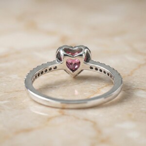 May include: A silver ring with a heart-shaped pink gemstone at the center. The band is adorned with small, clear stones. The ring is set against a light-colored, marble-like surface.