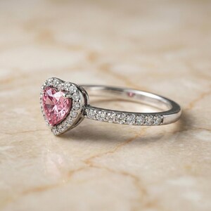 May include: A silver ring with a heart-shaped pink gemstone center, surrounded by a halo of smaller white gemstones. The band is also adorned with small white gemstones. The ring is set against a light-colored background.