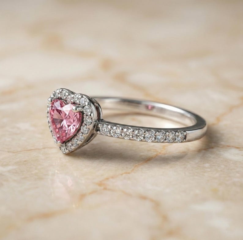 May include: A silver ring with a heart-shaped pink gemstone center, surrounded by a halo of smaller white gemstones. The band is also adorned with small white gemstones. The ring is set against a light-colored background.