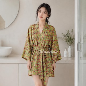 Könnte beinhalten: Ein kurzer Bademantel im Kimono-Stil mit einer leuchtend grünen Basis und einem sich wiederholenden Muster aus kleinen roten Blumen und dunkelbraunen Stielen. Das Kleidungsstück hat weite Ärmel, einen V-Ausschnitt und einen passenden Bindegürtel. Der Text ThePuranaBazar ist sichtbar.