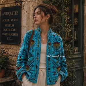 Pode incluir: Um casaco turquesa brilhante com gola alta e mangas compridas, adornado com bordados florais intrincados em tons de vermelho, verde e azul escuro. A peça tem uma abertura frontal reta e é usada sobre um top branco liso e calças claras.