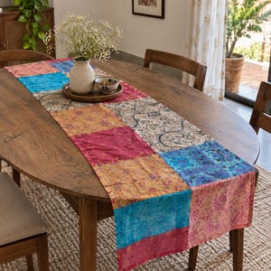 Könnte beinhalten: Ein rechteckiger Tischläufer mit einem Patchwork-Design aus verschiedenen Stoffquadraten in Blau-, Rosa-, Orange- und Cremetönen mit komplizierten Blumen- und Paisley-Mustern, flach ausgelegt in der Mitte eines ovalen hölzernen Esstisches.
