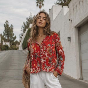 May include: A long-sleeved button-down shirt in a vibrant red fabric featuring a multi-coloured floral and botanical print, paired with loose-fitting white wide-leg trousers with side pockets and tan open-toe sandals, accompanied by a fringed bag.
