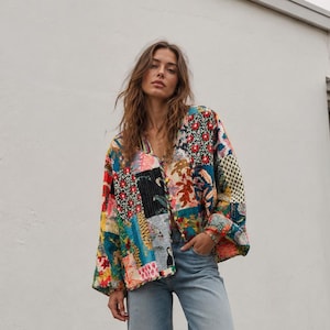 Op de afbeelding: Een jas met lange mouwen voorzien van een levendig patchworkontwerp met diverse bloemen- en geometrische patronen in rood, blauw, geel en groen, gecombineerd met een lichtblauwe spijkerbroek met wijde pijpen en een rafelige zoom.