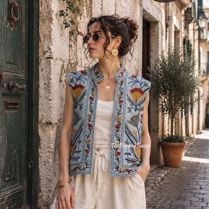May include: A light blue sleeveless waistcoat featuring intricate floral embroidery in shades of red, yellow, and dark blue. The garment has a high collar, a straight front opening, and is worn over a plain cream-coloured top and matching trousers.