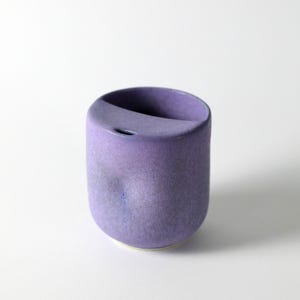 Può includere: Un vaso cilindrico in ceramica color lavanda con un'apertura superiore curva. Il vaso ha una finitura liscia e opaca e un piccolo foro vicino alla parte superiore. La base è di una tonalità più chiara di lavanda. Il design è minimalista e moderno.