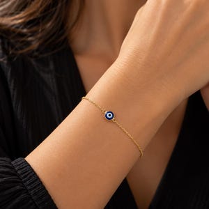 May include: A delicate gold chain bracelet featuring a single circular evil eye charm. The charm consists of a dark blue center surrounded by a white ring and a thin gold border, centered on a fine, polished gold-tone metal link chain.