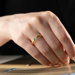 May include: A gold ring featuring a rectangular green gemstone. The ring is worn on a finger, with the hand in the foreground. The ring is the central focus, set against a blurred background.